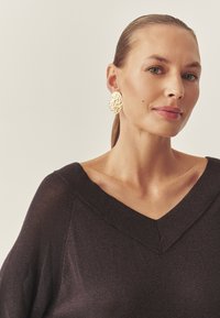 Fekete pulóver V-nyakkivágással, finom csillogással. Arany színű, texturált, ovális fülbevalók egészítik ki a megjelenést. Simára fésült, hátra kötött haj.