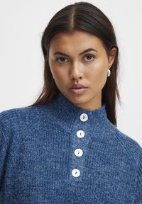 Pull en tricot bleu foncé avec un col haut et quatre grands boutons blancs. Présente un motif texturé et des détails côtelés.