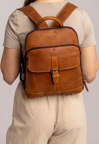 Mochila de cuero marrón con un diseño estructurado, bolsillo frontal, correas ajustables y un asa superior. Presenta detalles de costura y cremalleras.