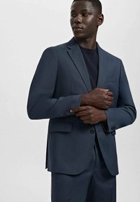 Mężczyzna poprawiający rękaw ciemnoniebieskiej marynarki, w dopasowanych spodniach i ciemnej koszuli, stojący na jednolitym jasnym tle.