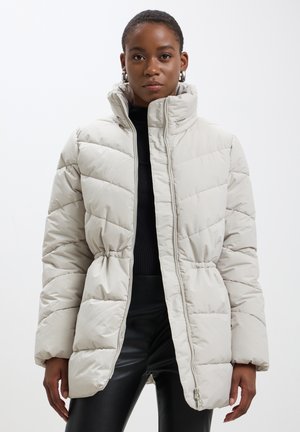 Veste matelassée beige clair avec un col haut, une fermeture éclair et une taille froncée. Présente des coutures horizontales et une texture lisse et brillante.