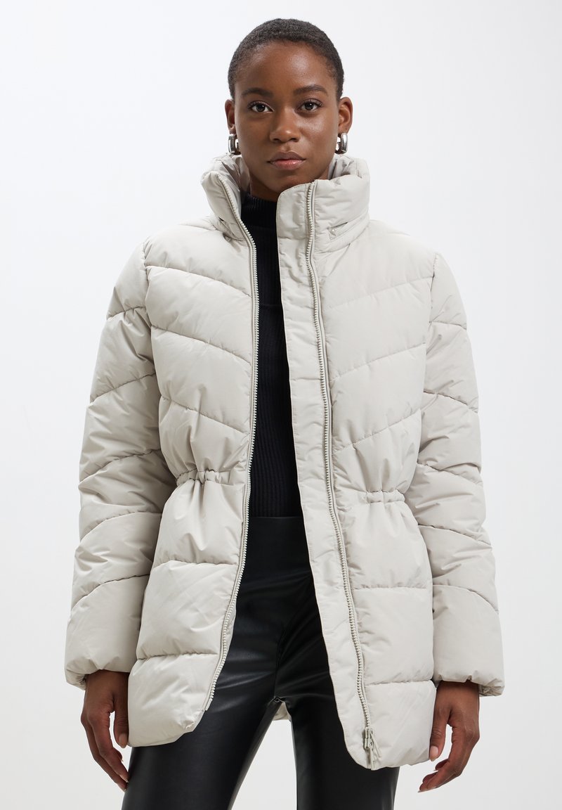 Veste matelassée beige clair avec un col haut, une fermeture éclair et une taille froncée. Présente des coutures horizontales et une texture lisse et brillante.