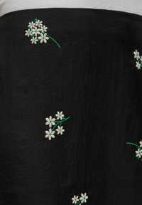 Schwarzer Stoff mit einer strukturierten Oberfläche, die mit weißen gestickten Blumen und grünen Stängeln verziert ist, die in einem verstreuten Muster angeordnet sind.