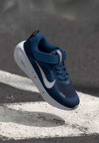 Sininen urheilukenkä, jossa on verkkopäällinen, valkoiset korostukset ja hihna. Varustettu teksturoidulla pohjalla ja erottuvalla swoosh-logolla. Kevyt muotoilu.