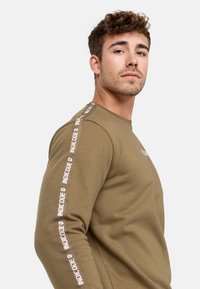 Olive green long-sleeve shirt made of cotton with white logo text on shoulder seams and a crew neckline. Soft texture, fitted design.