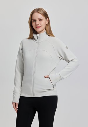 Ung kvinde med lige lyst blondt hår iført en lysegrå lynlåstrøje og sorte bukser, stående med hånden i lommen foran en ensfarvet baggrund.