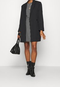 Oversized svart blazer lagd över en rutmönstrad klänning, i kombination med en svart handväska och knähöga svarta stövlar. Slät tyg, enkel design.