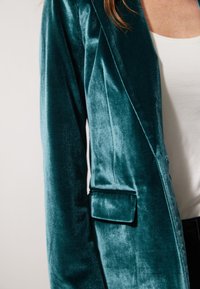 Teal velvet blazer with smooth texture, structured lapels, and a single pocket on the left side. Worn over a white top.