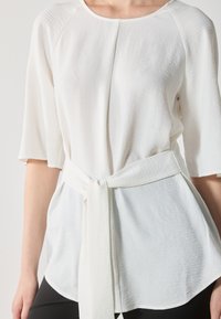 Blouse blanche à manches mi-longues, en tissu texturé, avec une ceinture à nouer à la taille. L'ourlet est arrondi, offrant une coupe décontractée.