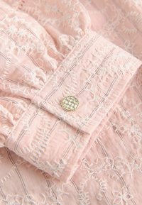 Tessuto rosa chiaro con motivi floreali ricamati bianchi e sottili righe nere, con un primo piano di un polsino piegato con un bottone argentato testurizzato.