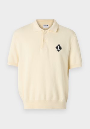 Koszulka polo w jasnym żółtym kolorze, z czarnym logo w kształcie diamentu na lewej piersi, dwoma guzikami przy kołnierzyku oraz krótkimi rękawami zakończonymi ściągaczami.