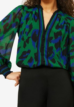 Kobieta w przezroczystej zielonej bluzce z niebiesko-czarnym abstrakcyjnym wzorem i czarnymi guzikami, w połączeniu z czarnymi spodniami z wysokim stanem.