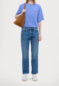 Vaaleansininen t-paita, jossa on punaista tekstiä, yhdistettynä suoriin sinisiin farkkuihin, valkoisiin lenkkareihin ja ruskeaan mokkanahkakelkkulaukkuun. Yksinkertainen, rento asukokonaisuus.