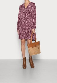 Robe fleurie bordeaux à manches longues, décolleté en V, et jupe froncée ; associée à un sac en cuir tan tissé et des bottines brunes.