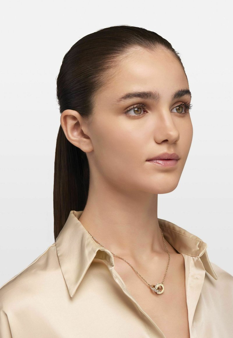 Goldkette mit einem runden Anhänger mit strukturiertem Design, getragen über einem Satinhemd in hellen Farben mit spitzem Kragen.