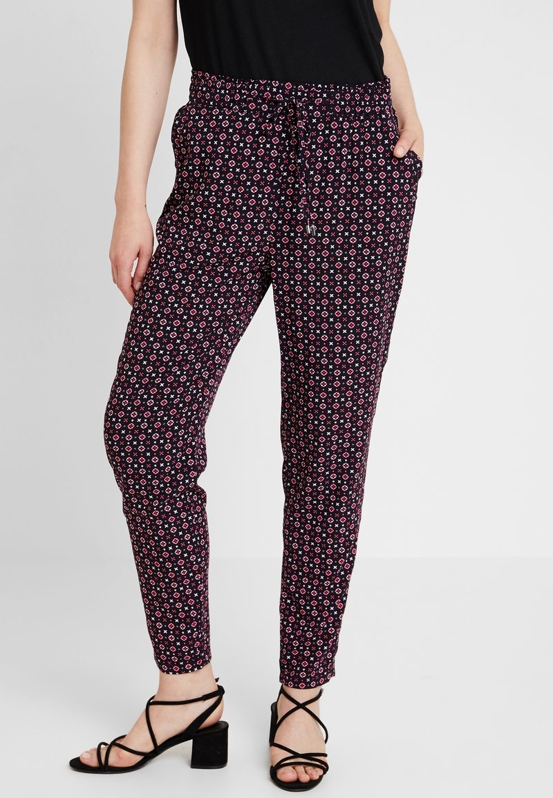 Femme portant un pantalon slim noir avec de petits motifs géométriques roses et blancs, associé à des sandales à talons blocs noires avec des lanières.