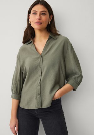 Vrouw draagt een licht olijfgroene blouse met knopen en driekwart mouwen, gecombineerd met een donkere spijkerbroek, staand tegen een effen lichte muur.