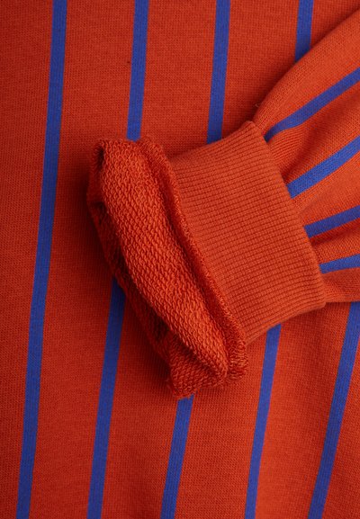 Tejido de punto rojo con rayas verticales azules. Presenta un puño texturizado en un rojo complementario. Acabado suave y agradable al tacto en general.