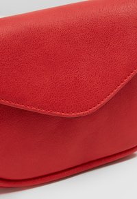 Punainen nahkainen pussukka, jossa on tekstuuripinta, kaareva läppä ja näkyvät ompeleet reunoilla. Kompakti ja tyylikäs muotoilu.