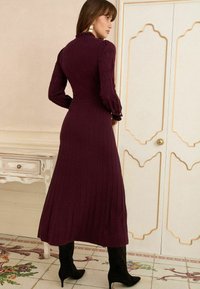 Duga, rebrasta, burgundska haljina dugih rukava s detaljem volančića na ovratniku. Nosena s crnim, špicastim čizmama do gležnja. Elegantni i uski dizajn.