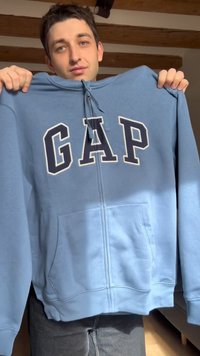 Joven sosteniendo una sudadera con capucha azul con cremallera y grandes letras "GAP" en el frente, en el interior de un lugar con vigas de madera en el techo.