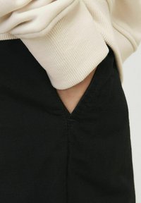 Beige pulóver mandzsettája fekete anyagon, amely oldalsó zsebet tartalmaz. A fekete anyag finom textúrájú, és látható varrás részletek találhatóak rajta.