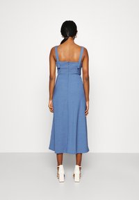 Mujer de pie de espaldas a la cámara, vestida con un vestido midi sin mangas azul y sandalias de tacón cuadrado blancas, frente a un fondo liso.