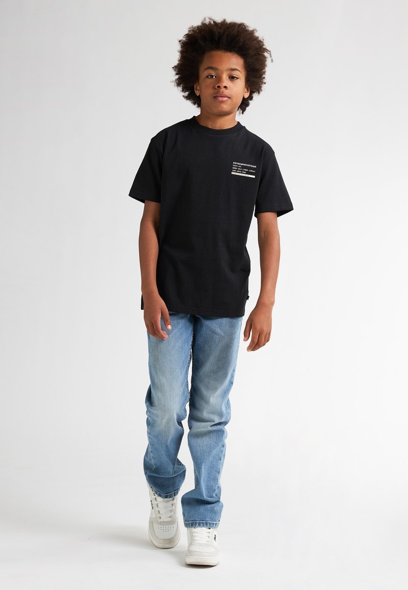 Zwarte t-shirt met korte mouwen en bedrukte tekst op de borst, gecombineerd met lichtblauwe jeans en witte sneakers, staand tegen een effen achtergrond.