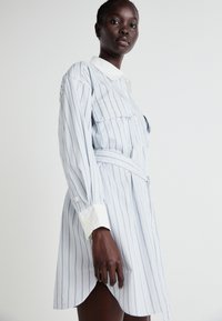 Krekli ar garām piedurknēm, gaiši ziliem un baltajiem svītrām, ar pieguļošu vidukli. Iekļauts balts apkakle un divas krūšu kabatas. Izgatavots no kokvilnas auduma.