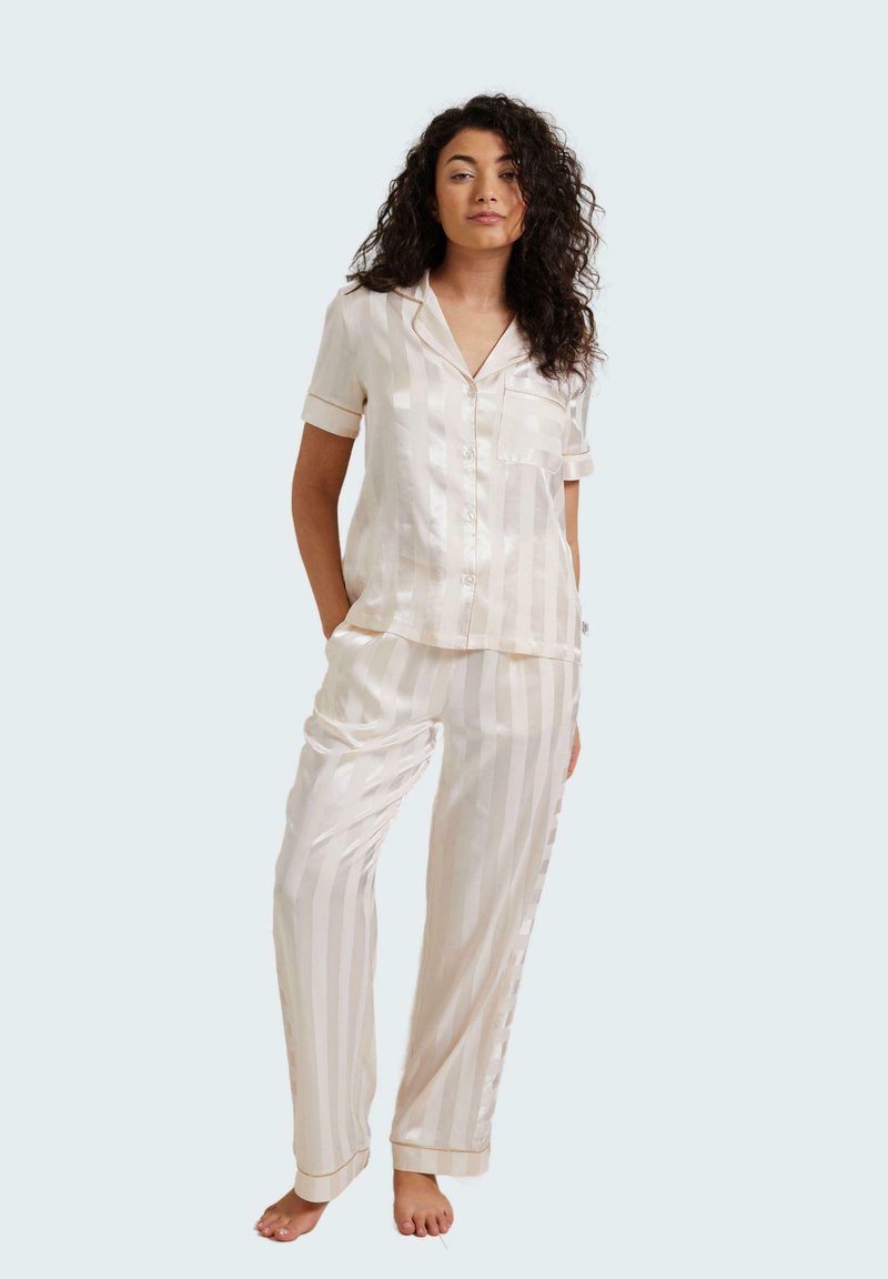 Light cream satin pajama set with striped pattern, featuring short-sleeved top with pocket and long, loose-fitting pants. Accented with trim.