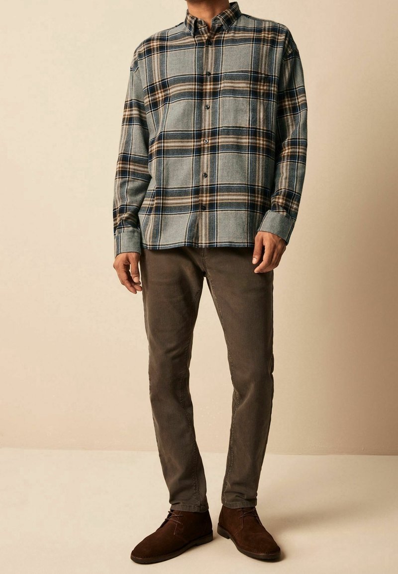 Homme portant une chemise à carreaux bleue, beige et noire boutonnée, un pantalon marron et des chaussures en daim marron foncé, debout devant un fond beige.