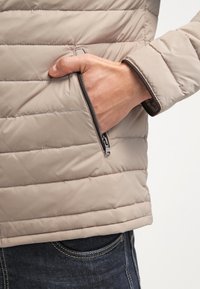 Doudoune beige avec coutures horizontales, dotée d'une poche zippée sur le côté gauche et d'un liseré marron foncé le long des bords.