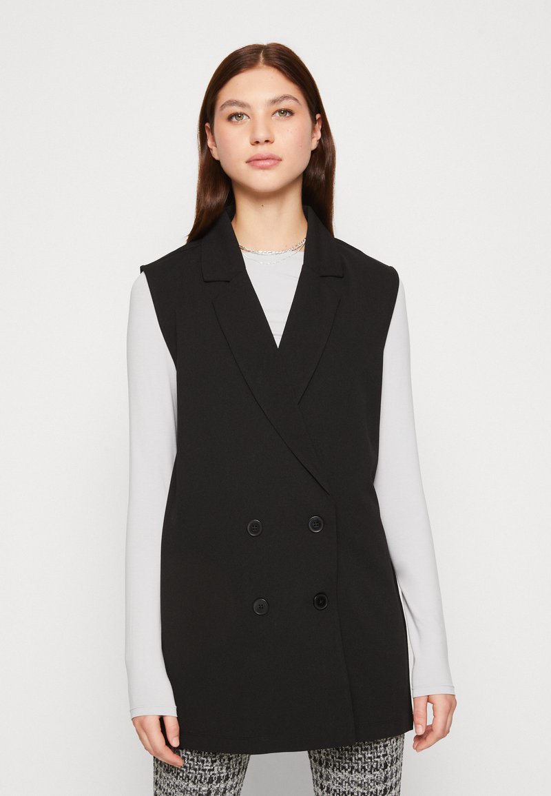 Blazer sin mangas negro con un diseño de doble botonadura, solapas con muesca y textura sutil. Lleva puesto una camiseta de manga larga en gris claro.