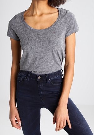 Donna con t-shirt grigia aderente con scollo a barca e jeans skinny blu scuro a vita alta, in piedi davanti a uno sfondo semplice.