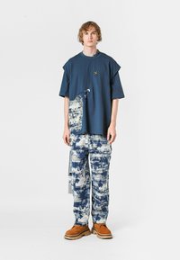 Joven vistiendo una camisa oversized de color azul marino con un diseño de pintura abstracta y pantalones a juego de patrón azul y blanco, con zapatos marrones.