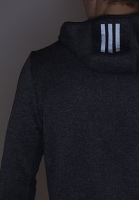 Sweat à capuche gris foncé avec un col montant et trois bandes blanches sur l'arrière de la capuche, fabriqué en tissu texturé.