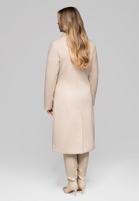 Mujer con cabello largo y rubio lleva un abrigo beige hasta la rodilla y botas de tacón a juego, de pie frente a un fondo gris liso.