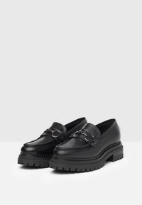 Mocassins en cuir noir avec une semelle en caoutchouc épaisse, dotés d'une boucle en métal décorative et de détails de couture élégants. Texture lisse et forme arrondie.