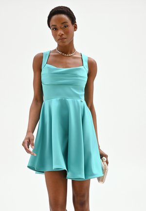 Donna che indossa un vestito azzurro senza maniche, con una pochette di perline, accessoriata con una collana a catena d'oro, su sfondo bianco semplice.