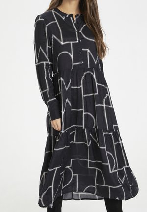 Femme portant une robe noire ample avec un motif de lettres abstraites blanches brodées et des manches longues sur un fond blanc uni.