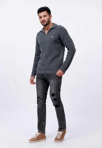 Moški v sivem puloverju s četrtim zadrgo, črnih raztrganih kavbojkah in rjavih supergah, stoji z eno roko v žepu ob enobarvni podlagi.