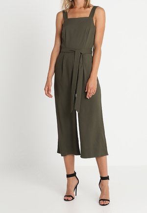 Femme portant une combinaison sans manches vert olive à jambes larges avec ceinture à la taille, associée à des sandales noires à talons hauts avec bride à la cheville.