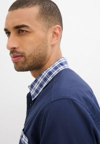 Camicia navy con colletto a quadri blu e bianco. Presenta una tasca frontale e cuciture a contrasto. Tessuto liscio, vestibilità sartoriale.