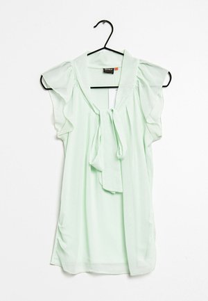 Blusa verde claro hecha de una tela transparente. Presenta un escote con lazo y mangas cortas con volantes. Cuelga de una percha de metal negro.