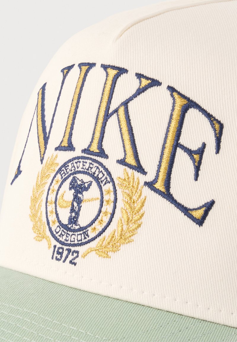Gorra blanca con visera verde bordada con el texto grande "NIKE" en azul y amarillo, emblema circular que presenta una figura alada, hojas de laurel y el texto "1972".