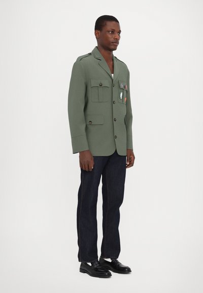 Veste cintrée vert olive avec deux poches avant et des patches décoratifs, associée à un jean en denim foncé et des mocassins noirs.