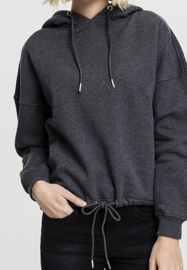 Personne portant un sweat à capuche gris foncé avec cordons à l'ourlet et pantalon noir, main partiellement dans la poche, sur un fond clair uni.