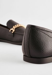 Black leather loafers with a textured finish, featuring a gold chain accent across the vamp and a rounded heel.