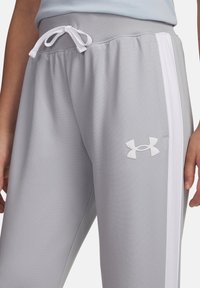 Šedé športové nohavice s bielymi bočnými pruhmi, so zaväzovaním v páse a logom Under Armour na pravej stehne, ktoré nosí stojaci človek.