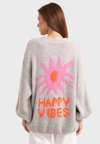 Grosse pullover gris avec un tricot texturé, arborant une grande fleur rose et un graphique orange "HAPPY VIBES" au dos.